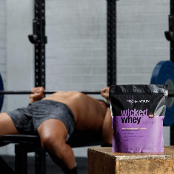 WICKED WHEY WHEY PROTEIN POWDERS 2kg CHOCOLATE, Isolate WICKED WHEY. Man in gym in background lifting weights. In foreground packet of wicked whey on bench