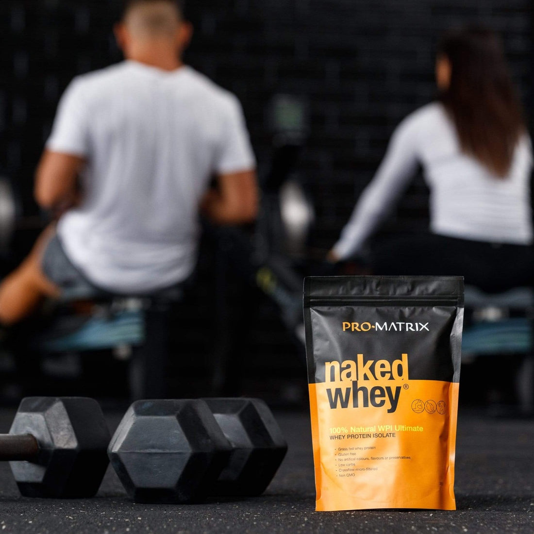 NAKED WHEY WHEY PROTEIN POWDERS 1kg UNFLAVOURED, Concentrate NAKED WHEY. Male and Female blurred out in background at gym on rowwers with their backs to camera. naked whey packet in foreground with two sets of hand weights