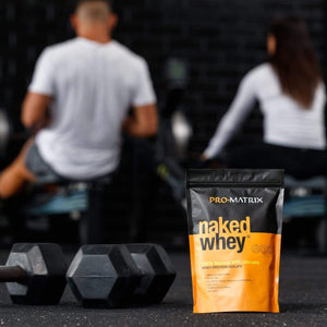 NAKED WHEY WHEY PROTEIN POWDERS 1kg UNFLAVOURED, Concentrate NAKED WHEY. Male and Female blurred out in background at gym on rowwers with their backs to camera. naked whey packet in foreground with two sets of hand weights