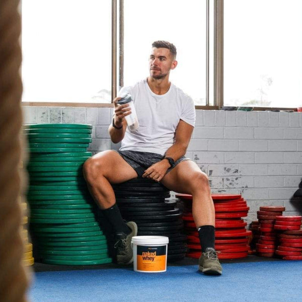 NAKED WHEY WHEY PROTEIN POWDERS 2kg UNFLAVOURED, Concentrate NAKED WHEY. Man in gym sitting on weights holding pro matrix shaker with bucket of naked whey on ground