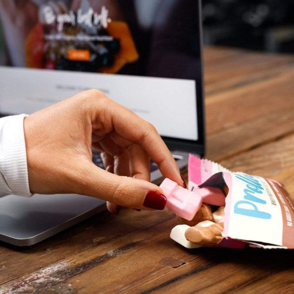 PROLLIES PROTEIN SNACKS PROLLIES - Whey protein lollies (BOX OF 10) Neapolitan Flavour. Person at desk with computer in background picking up prollies with open packet on bench. Pink prollie in shape of dumbbell