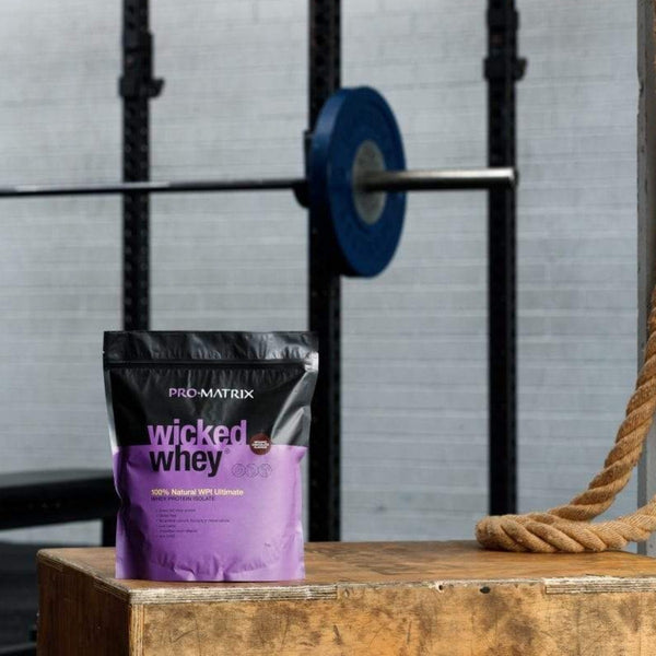 WICKED WHEY WHEY PROTEIN POWDERS 1kg CHOCOLATE, Concentrate WICKED WHEY. Wicked whey packet sitting on box in foreground with weight bar in background and training rope